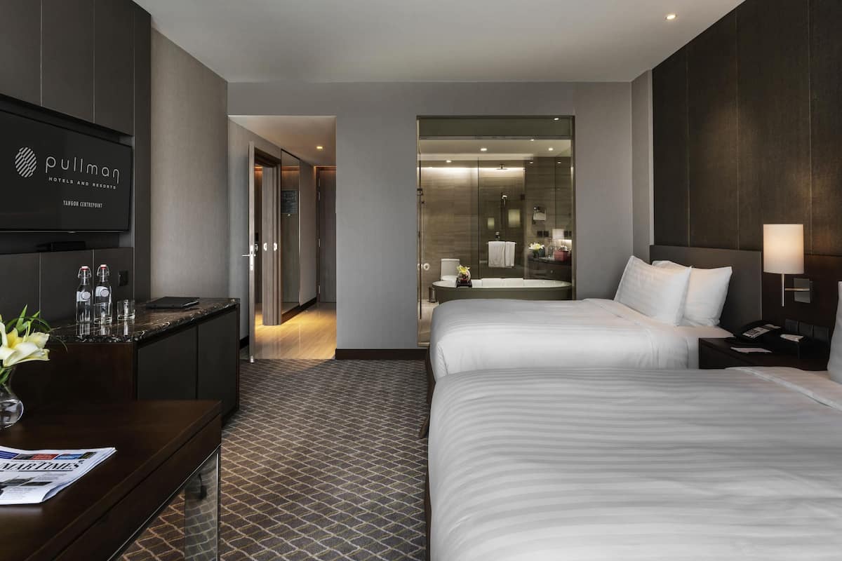 Ein Hotelzimmer mit zwei Betten, einem Schreibtisch mit Computer, einem Fernseher und einem durch eine Glastür einsehbaren Badezimmer.
