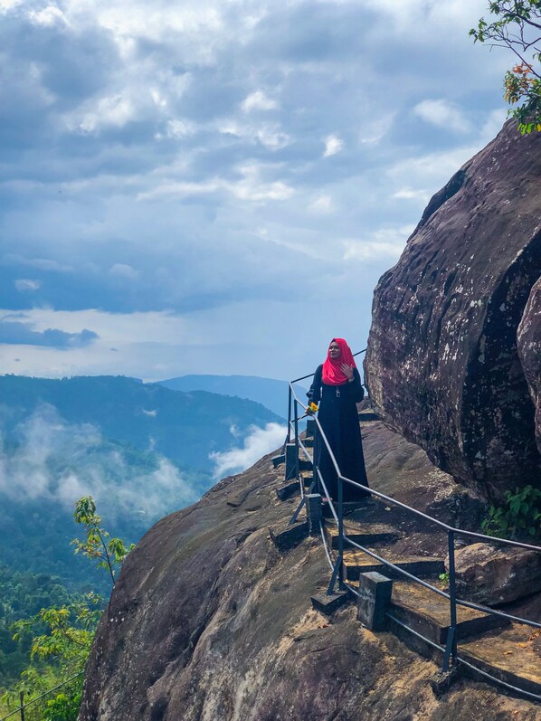 A person standing on a rocky outcrop with a metal railing, overlooking a valley with greenery and clouds below. - Misty Hills Villa Kadugannawa (Udunuwara)