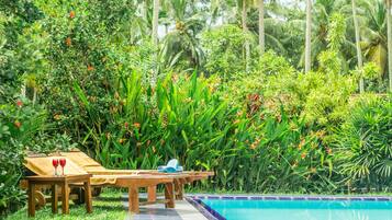A poolside area with a wooden table and lounge chairs, surrounded by lush greenery and palm trees.