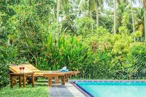 Zona junto a la piscina con mesa de madera y sillas reclinables, rodeada de abundante vegetación y palmeras.