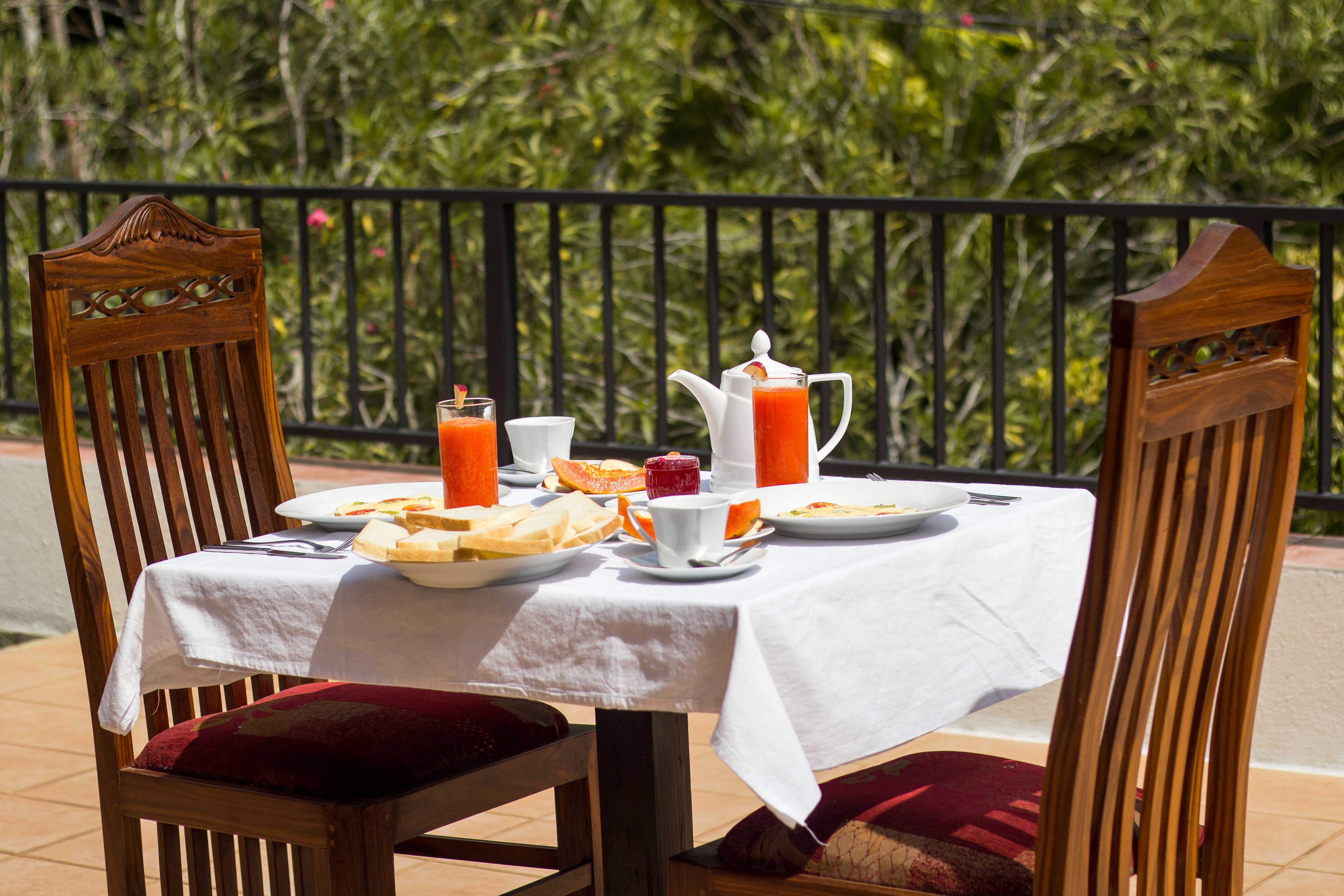 A table set for breakfast with a white tablecloth, wooden chairs, and a view of greenery in the background.