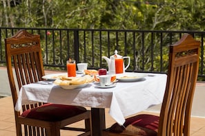 Mesa lista para el desayuno, con mantel blanco, sillas de madera y vista a la vegetación de fondo.