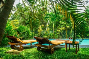 A poolside area with wooden lounge chairs and a table, surrounded by lush greenery and palm trees. - Sakura Villa (Bentota)