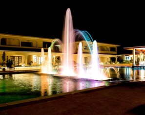 Una fuente nocturna con chorros de agua iluminados en varios colores, frente a un edificio con múltiples balcones y una piscina.