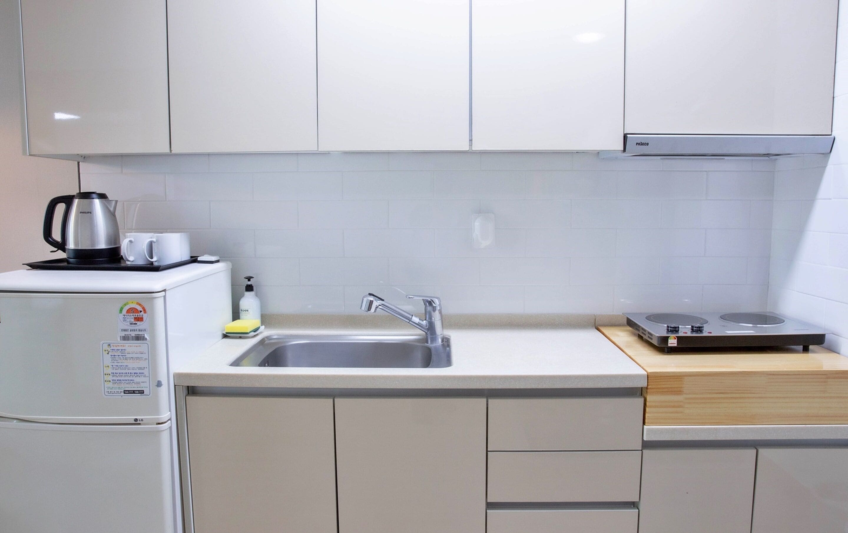 Cocina compacta con armarios blancos, pileta de acero inoxidable, cafetera, una botella de jabón para manos y una cocina portátil por inducción.