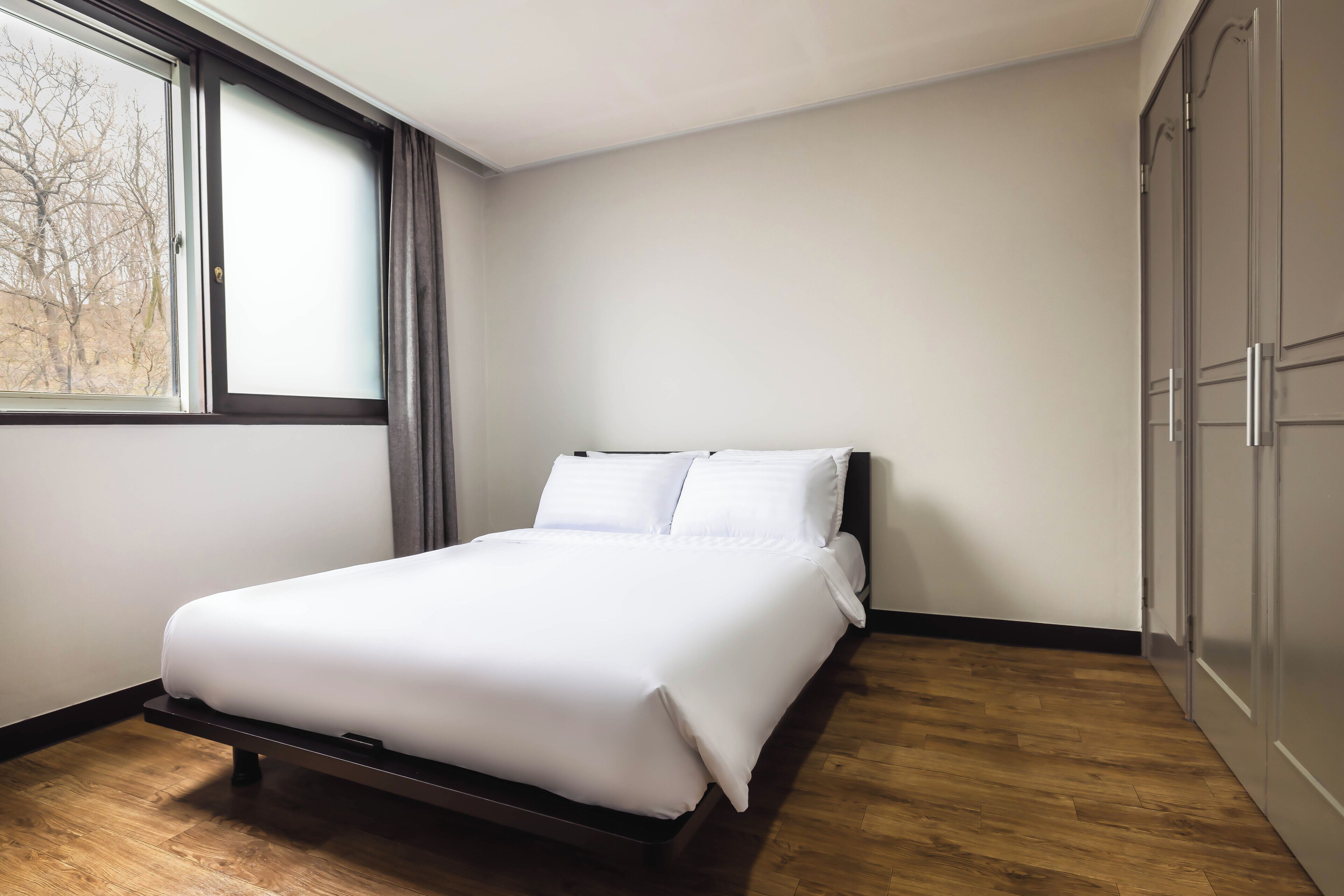 Cama bem arrumada em quarto com janela grande, piso de madeira e guarda-roupa.