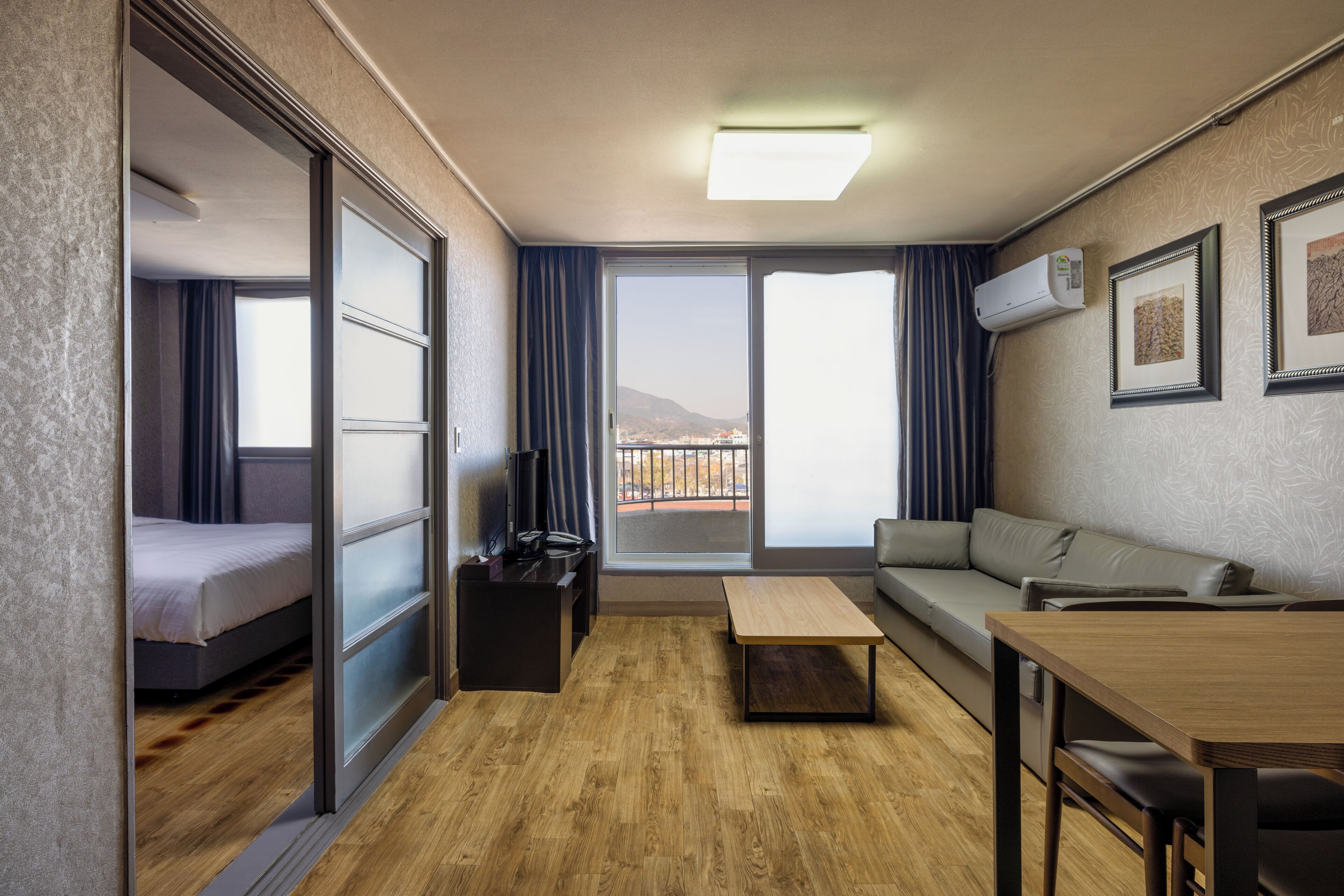 A modern hotel room with a bed, a sofa, a dining table, and a view of the city through large windows.