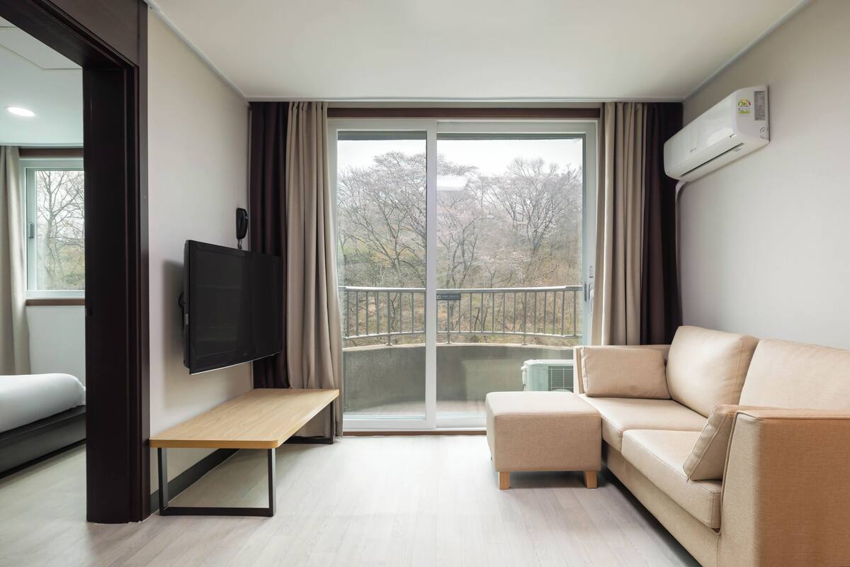 Un salon moderne avec un canapé, un téléviseur, un banc et un balcon offrant une vue sur les arbres.
