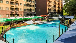 Une piscine dotée de parasols, d’un toboggan aquatique et offrant une vue sur un immeuble à plusieurs étages avec des balcons.