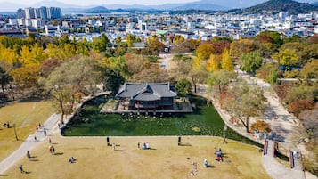 Un pabellón coreano tradicional rodeado por un estanque, con gente caminando y un paisaje urbano al fondo.