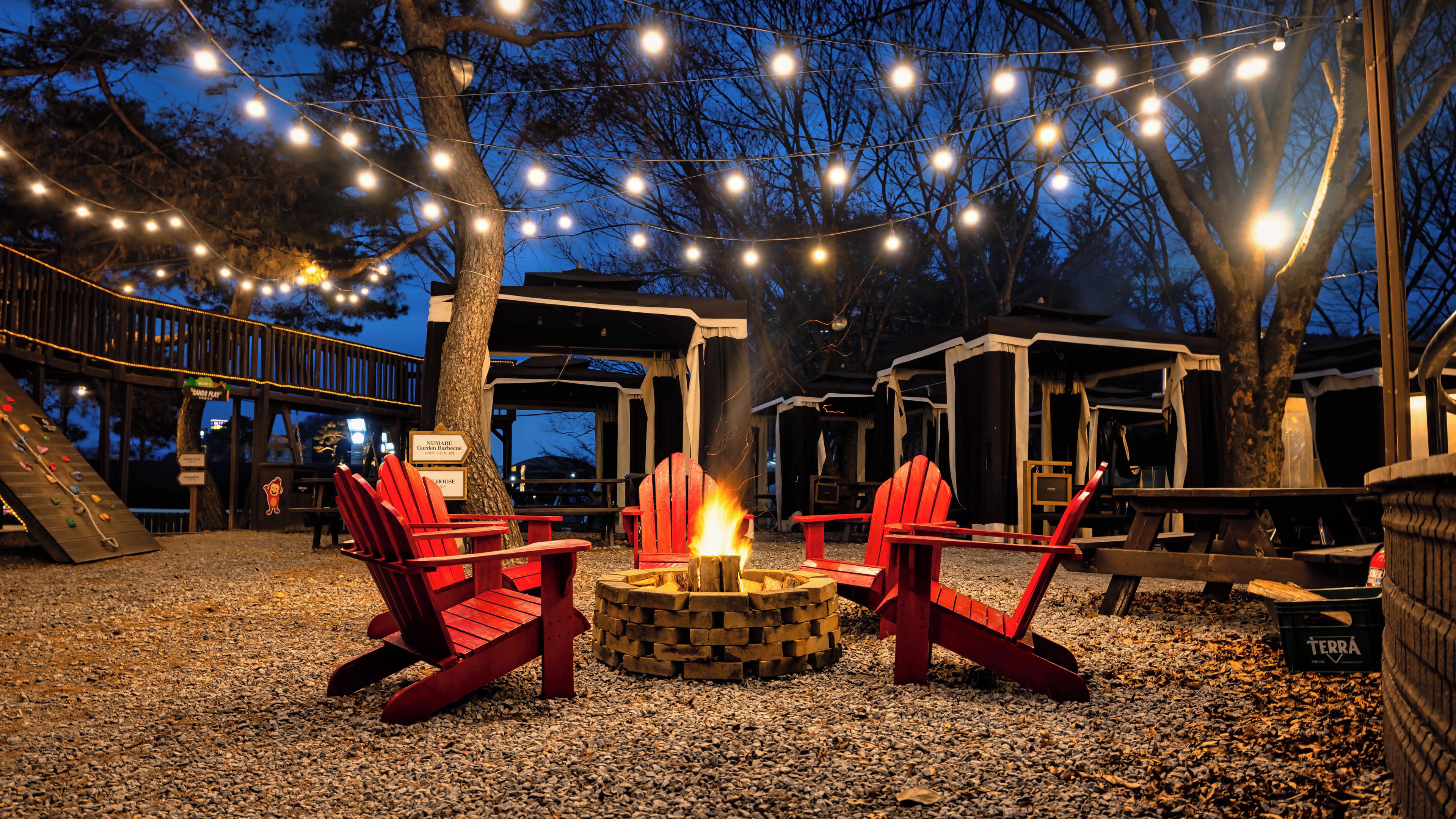 A nighttime outdoor seating area with red Adirondack chairs around a fire pit, string lights overhead, and a wooden deck.