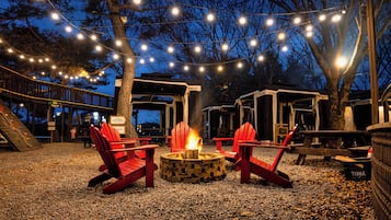A nighttime outdoor seating area with red Adirondack chairs around a fire pit, string lights overhead, and a wooden deck.