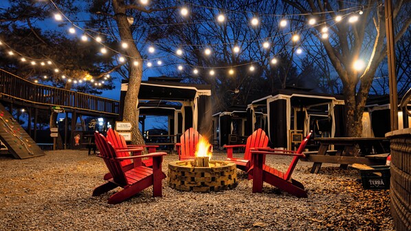 Un espacio al aire libre con asientos de noche, sillas Adirondack rojas alrededor de un brasero, luces colgantes y una terraza de madera.