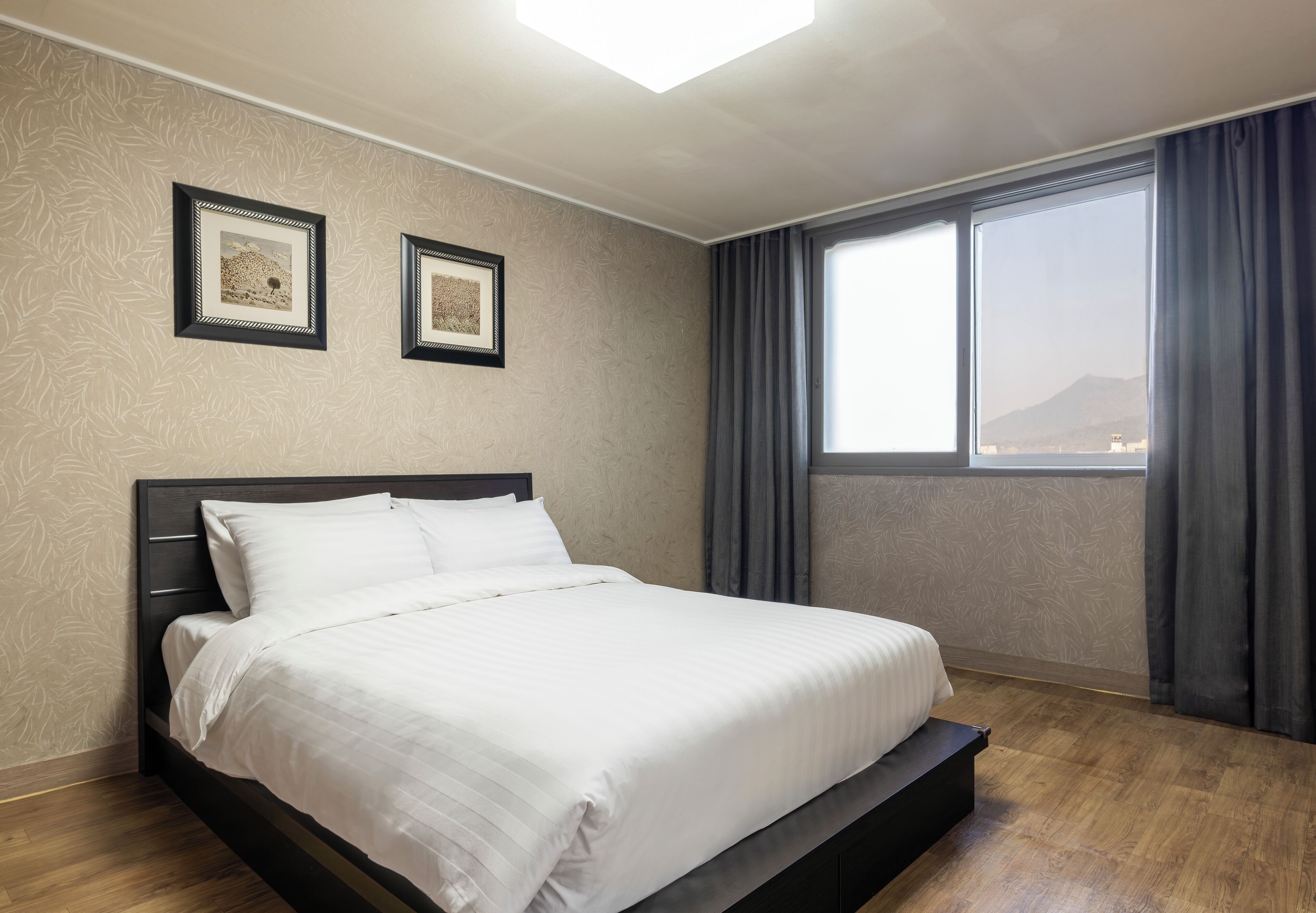 Una cama bien hecha en una habitación de hotel, con un ventanal grande, una lámpara de techo y dos cuadros en la pared.