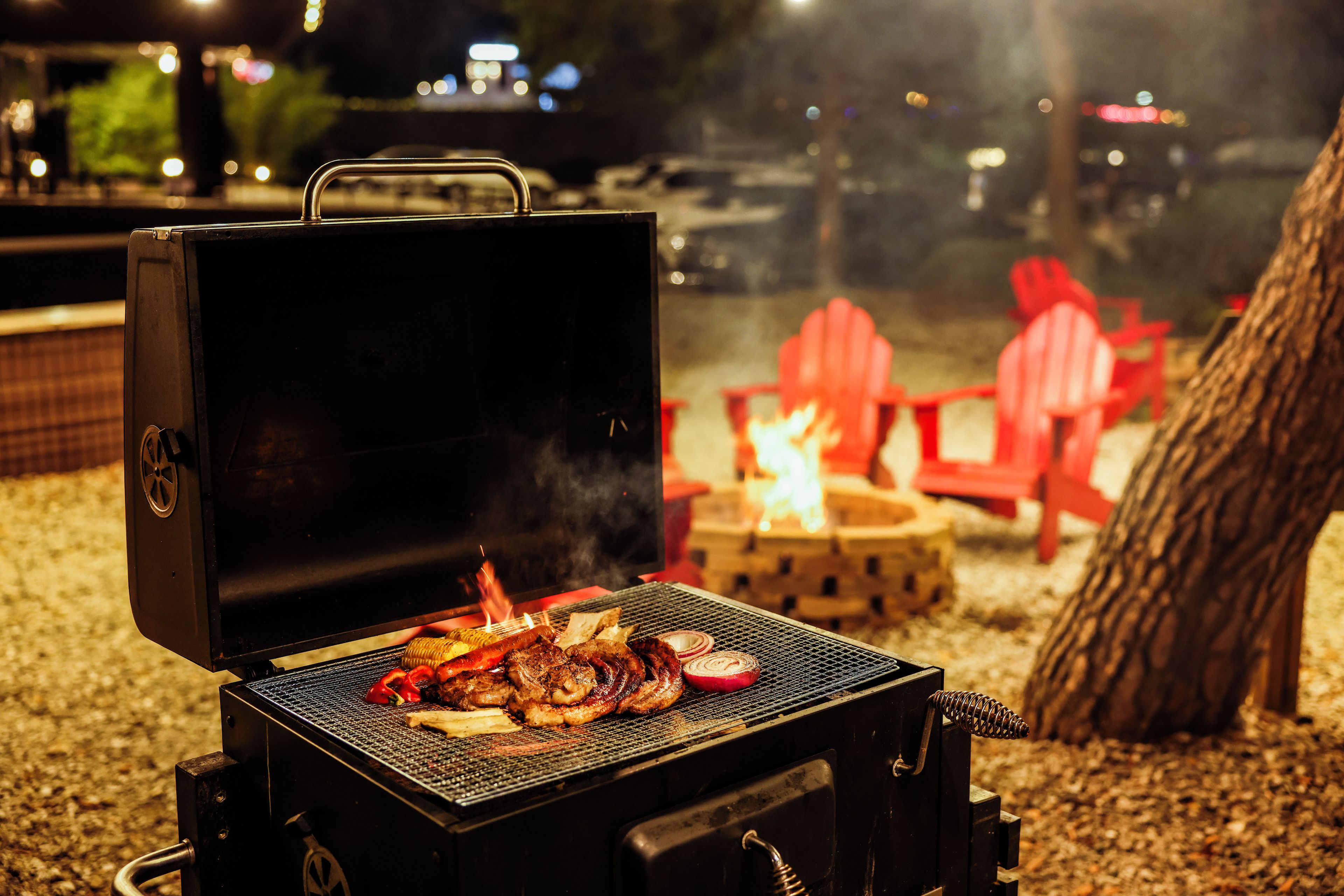 A barbecue grill with food being cooked, a fire pit, and red chairs in a nighttime outdoor setting.