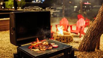 A barbecue grill with food being cooked, a fire pit, and red chairs in a nighttime outdoor setting.