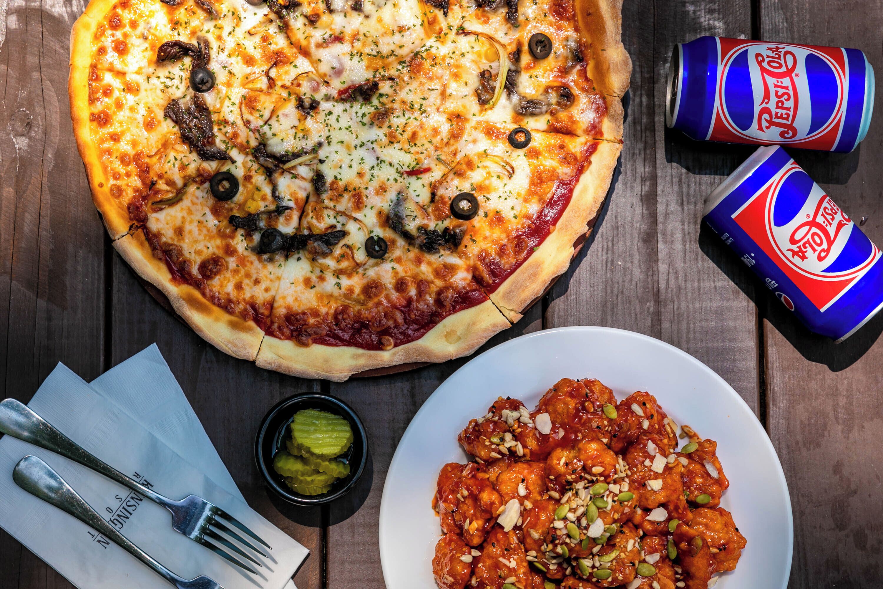 A pizza, a plate of chicken wings, and two cans of Pepsi on a wooden table.
