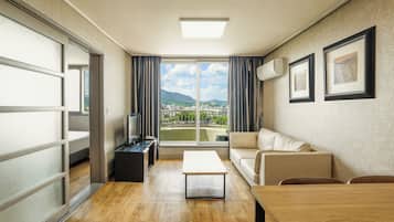 A modern hotel room with a sofa, a coffee table, a TV, and a view of a cityscape through large windows.