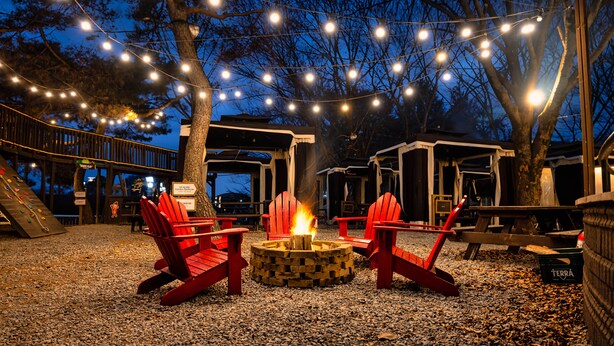 Un espace extérieur aménagé pour les soirées, avec des fauteuils Adirondack rouges disposés autour d’un foyer, des guirlandes lumineuses suspendues au-dessus et une terrasse en bois.