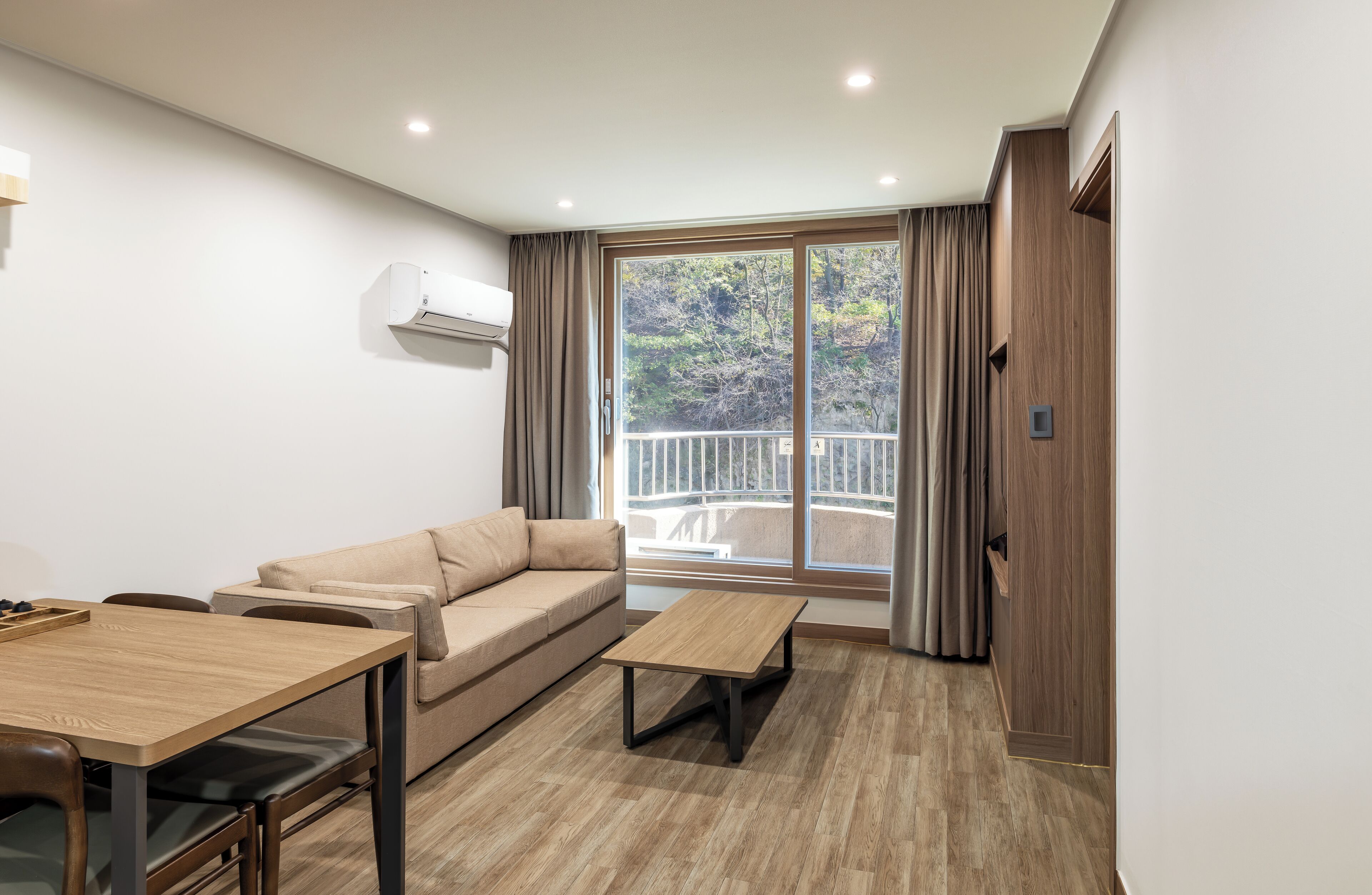 A modern living room with a sofa, a dining table, and a balcony with a view of trees.