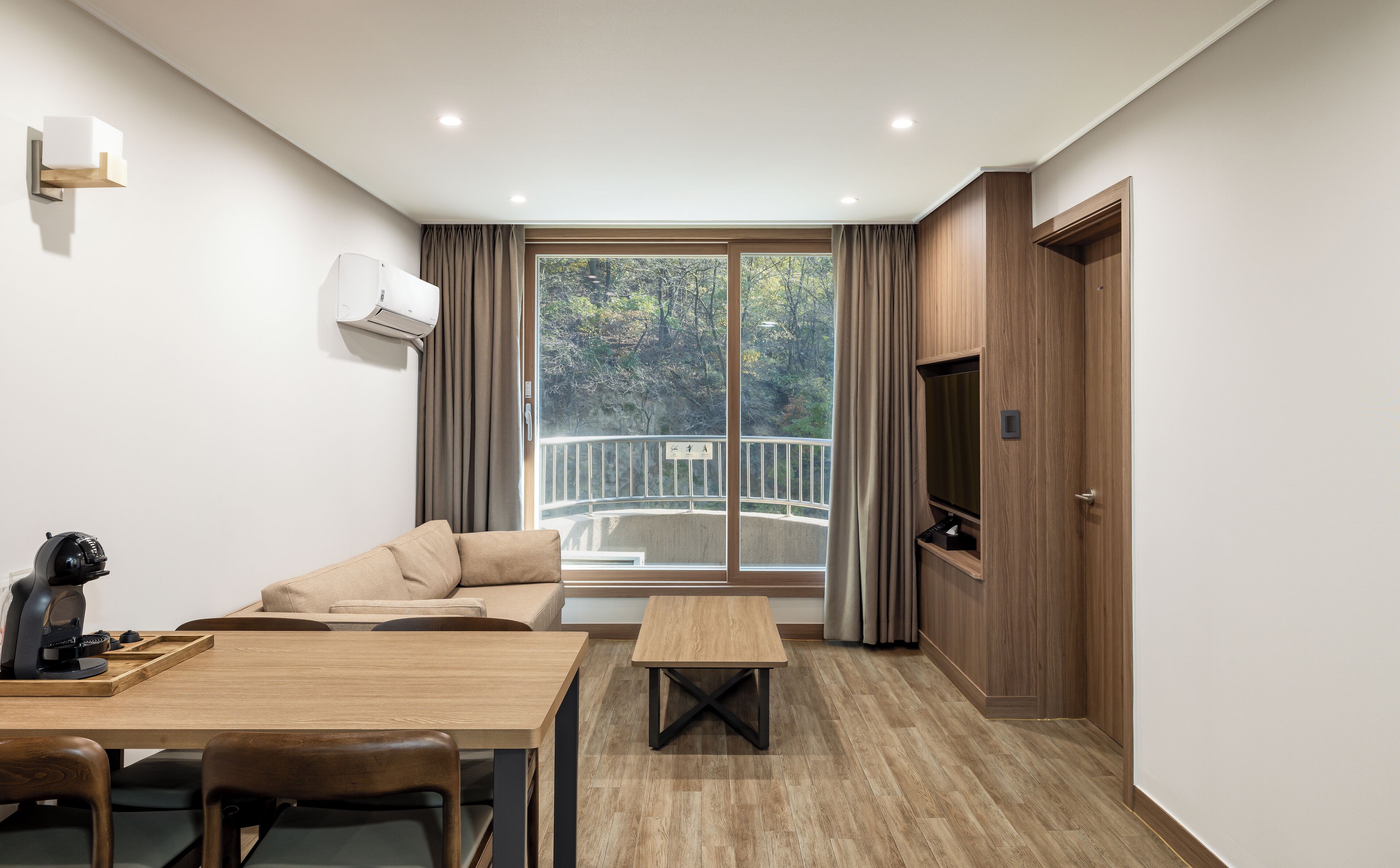 Habitación de hotel moderna con sofá, una mesa de madera, una cafetera y vistas a un balcón con barandilla.