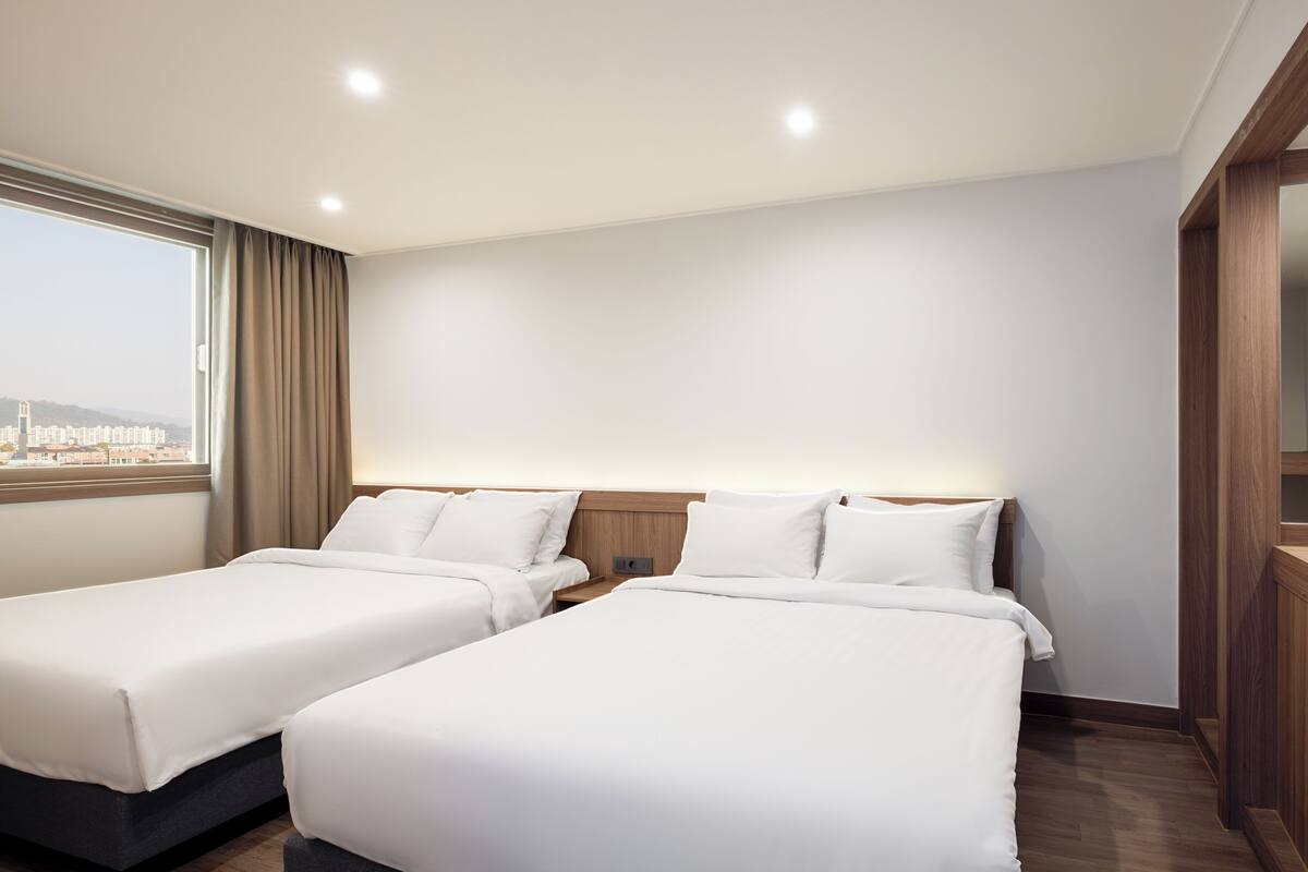 Une chambre d’hôtel avec deux lits, une grande fenêtre avec des rideaux, une tête de lit en bois et un miroir.