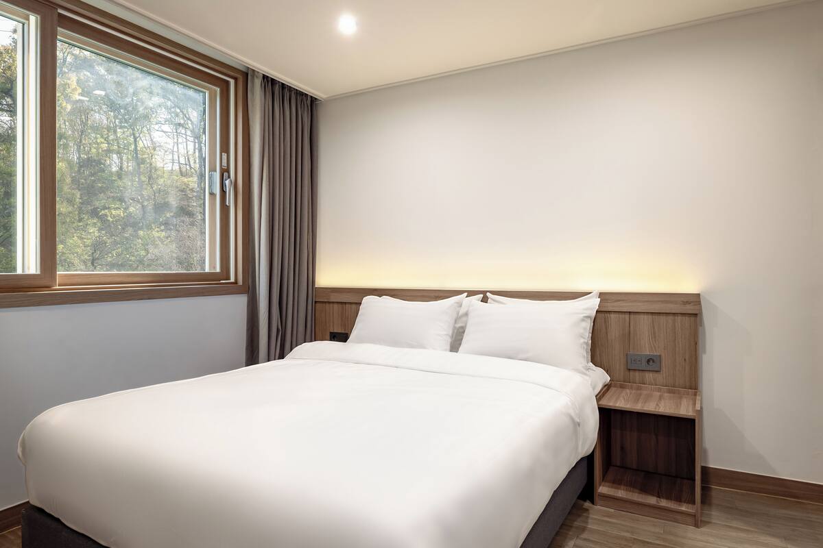 Un lit bien fait, avec du linge de lit blanc, dans une pièce dotée d’une grande fenêtre, d’une tête de lit en bois et d’une table de chevet.