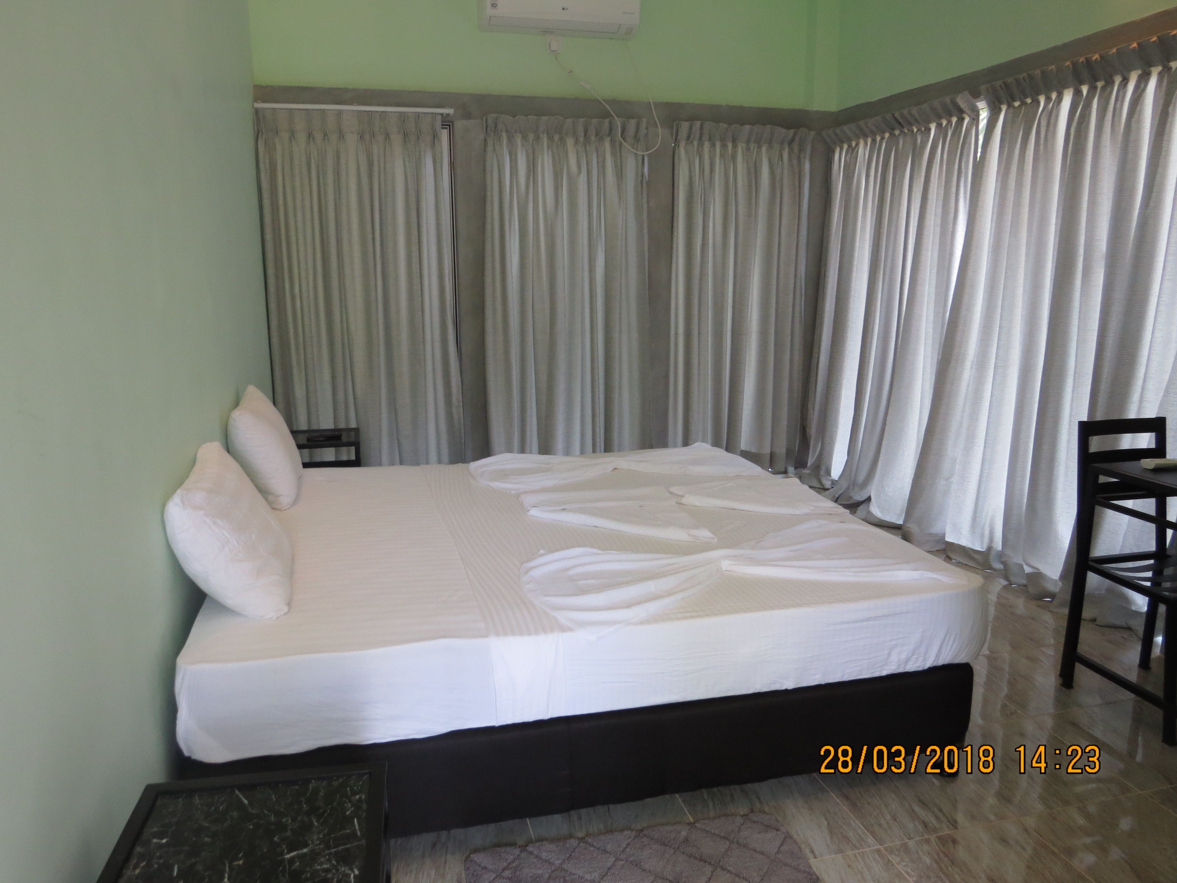 A neatly made bed in a room with a green wall, a black headboard, and a window with grey curtains. There is a small table and a chair to the right.