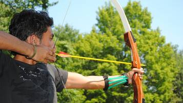 A person aiming a bow with a yellow arrow, wearing a black shirt with white text, outdoors with trees in the background.