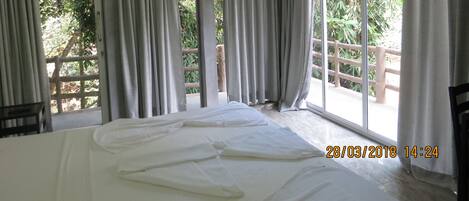 Quarto com cama, janelas amplas com cortinas, ar condicionado e vista para o verde lá fora.