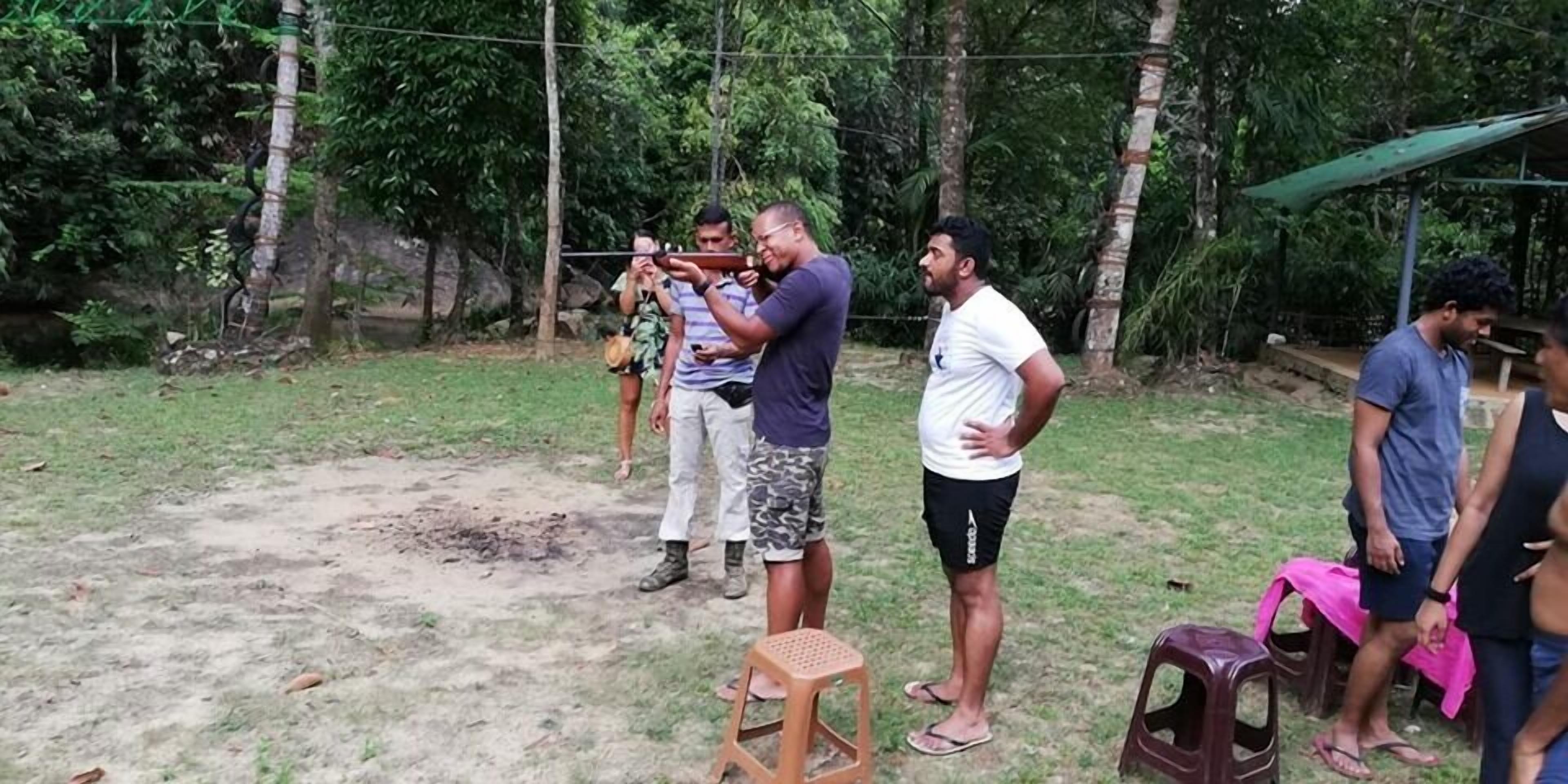 Un grupo de personas al aire libre, algunas de pie y otras sentadas en taburetes de plástico, con suelo de tierra y vegetación al fondo.