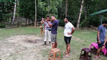 Un grupo de personas al aire libre, algunas de pie y otras sentadas en taburetes de plástico, con suelo de tierra y vegetación al fondo.