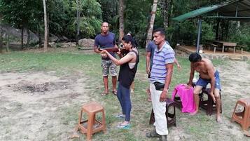 A group of people outdoors, some standing and some sitting on stools, in a grassy area with trees and a covered structure in the background.