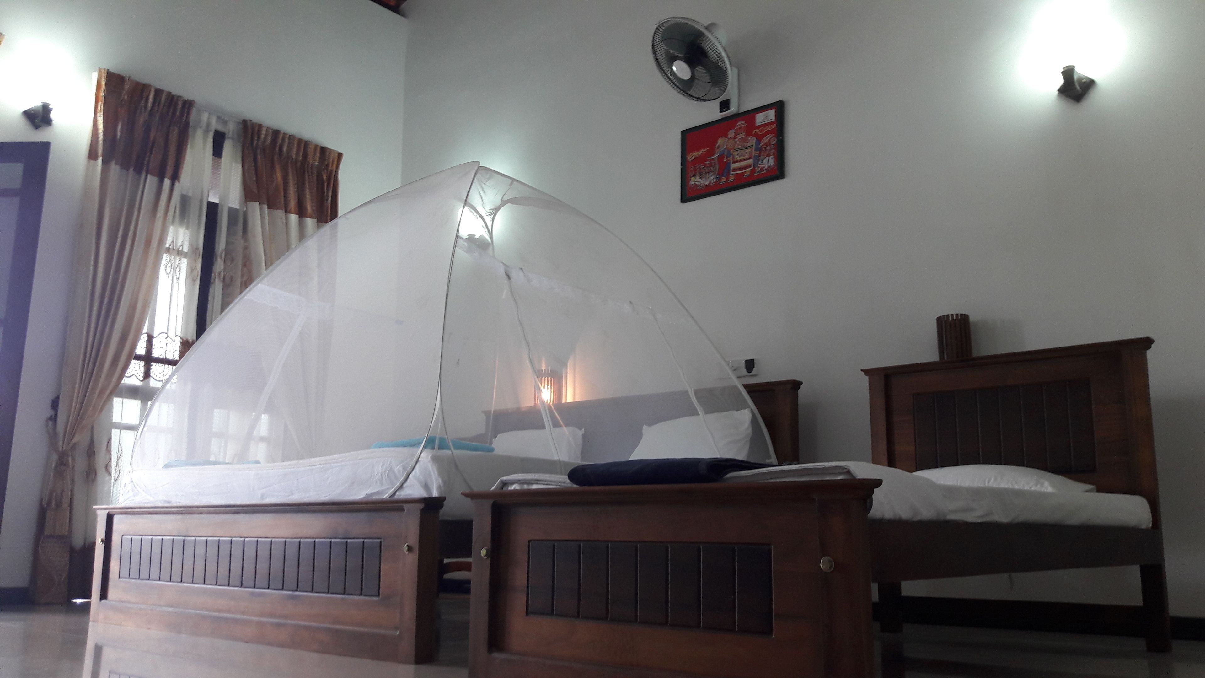 Un dormitorio con dos camas de madera, una con mosquitero. Hay un ventilador montado en la pared y un cuadro enmarcado sobre las camas.