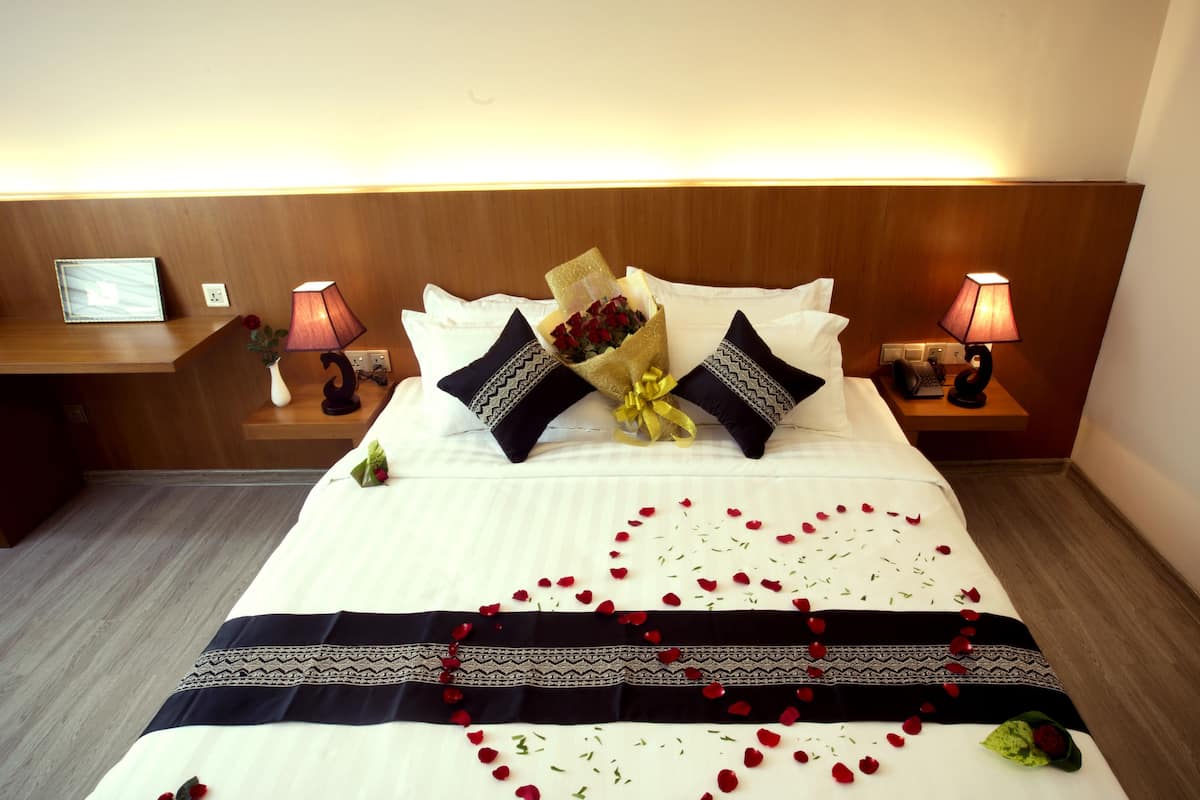 A neatly made bed with a heart-shaped arrangement of rose petals, two decorative pillows, and a bouquet of flowers.