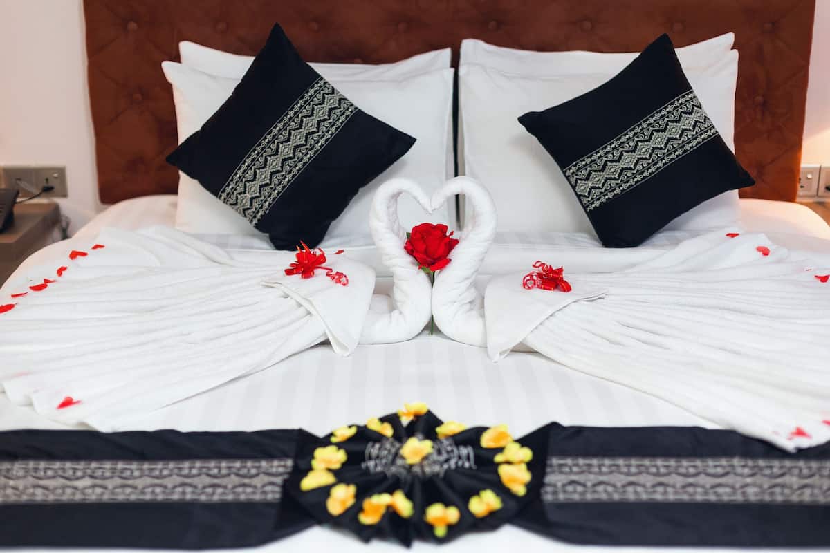 A neatly made bed with white linens, two black pillows with patterned trim, and a decorative arrangement of white towels and red roses forming a heart shape.
