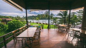 Une terrasse extérieure en bois, dotée de tables et de chaises, qui offre une vue sur une étendue d’eau et des palmiers.