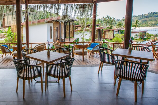 An outdoor patio with wooden tables and chairs, a view of the beach, and a small building in the background.
