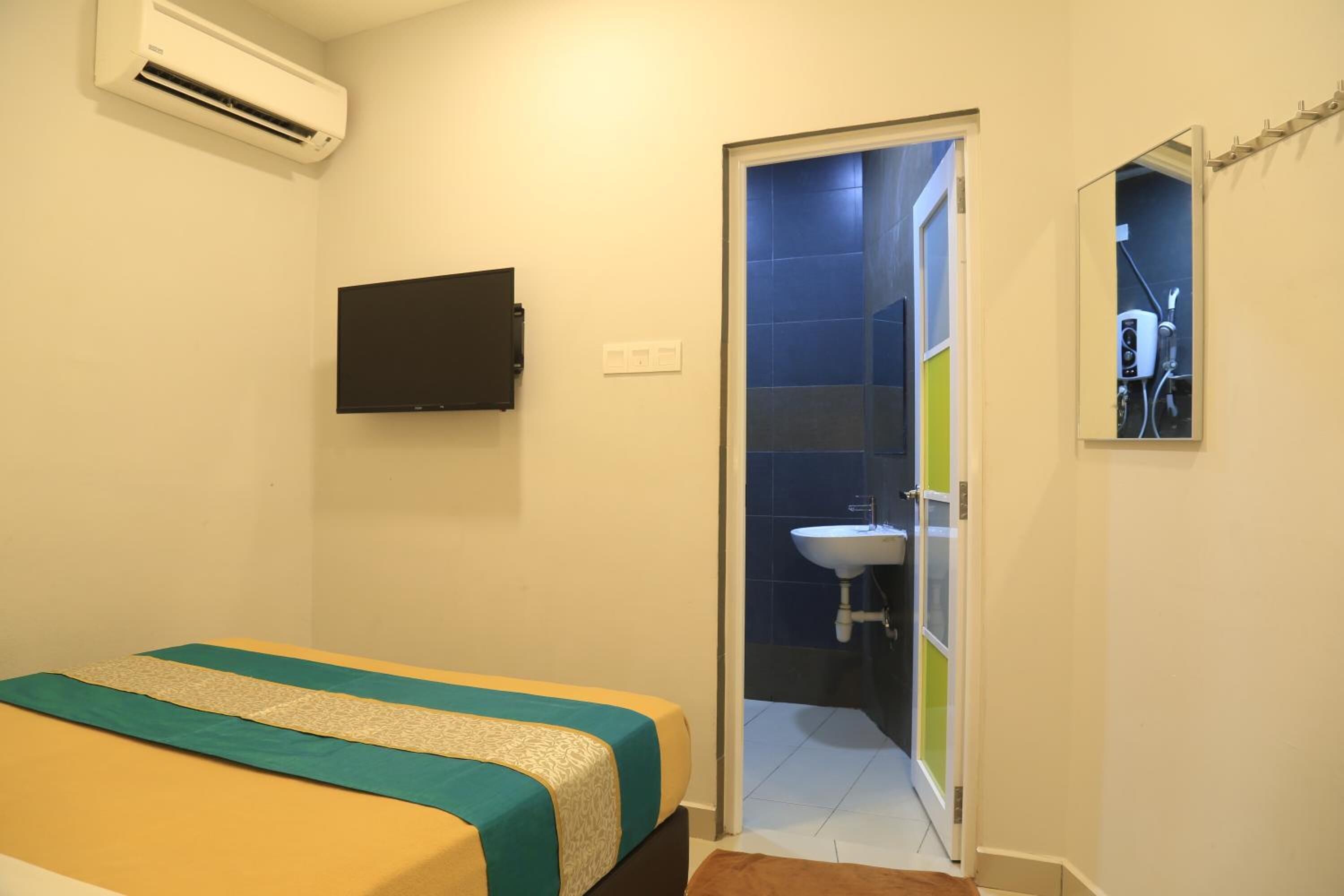 Una habitación con cama, televisor de pantalla plana, aire acondicionado, baño con lavamanos y espejo, y una puerta que da a otra habitación.