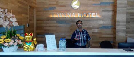Balcão de recepção com parede de madeira, uma placa com a inscrição "LAVINA HOTEL", uma estatueta dourada de gato, um arranjo de flores e um calendário.
