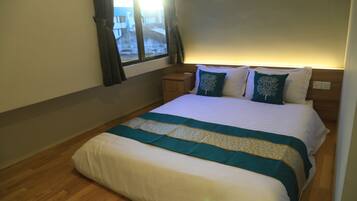 Un lit bien fait, recouvert d’une couette blanche et turquoise, une table de chevet en bois avec une lampe, et une fenêtre avec des rideaux.