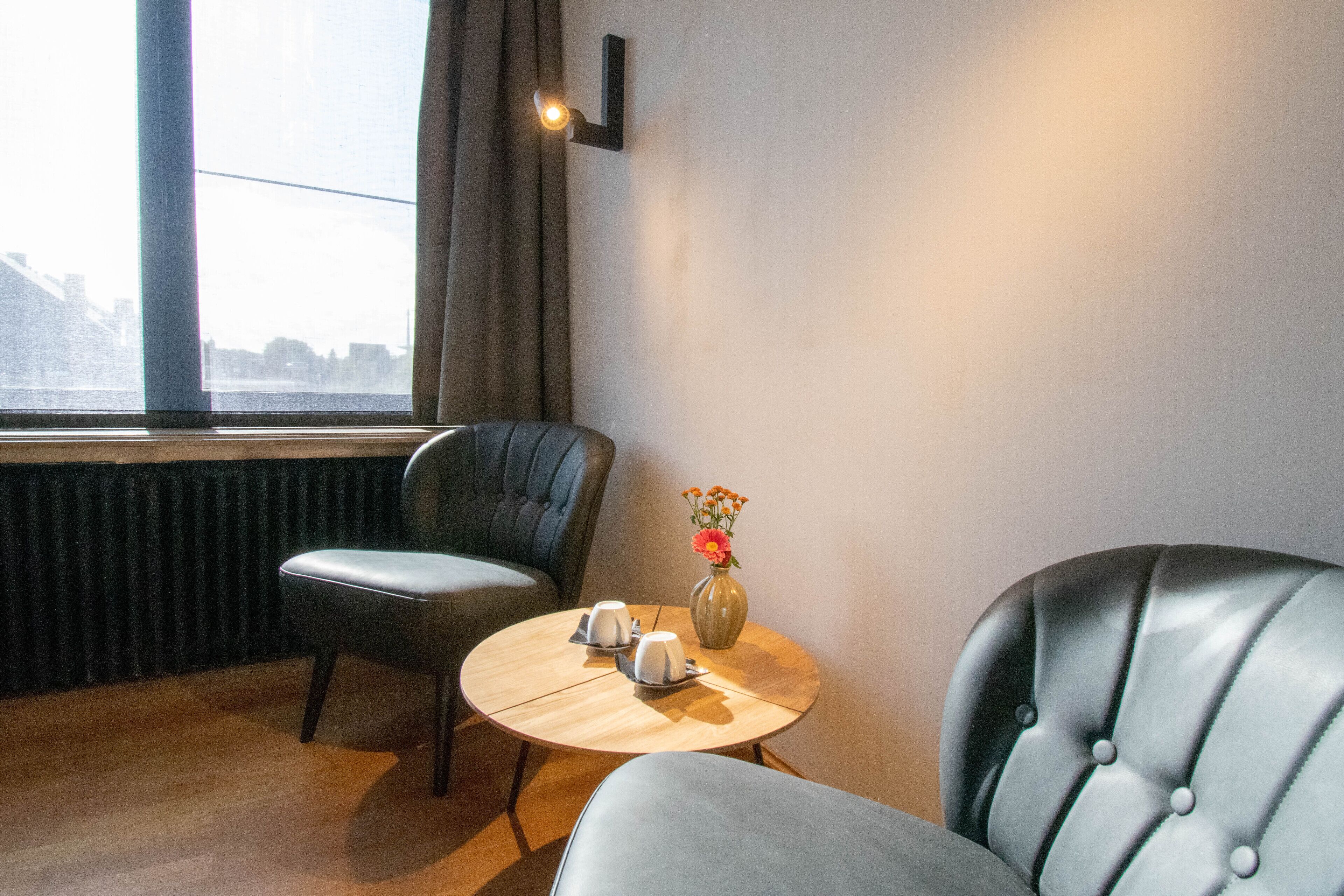 Une chambre d’hôtel avec vue sur le paysage urbain, visible à travers une grande fenêtre, une table ronde en bois avec deux tasses et un vase de fleurs, et deux fauteuils rembourrés.
