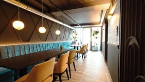 Un restaurante moderno con una larga mesa de madera, asientos acolchados, iluminación colgante y un diseño geométrico en la pared.