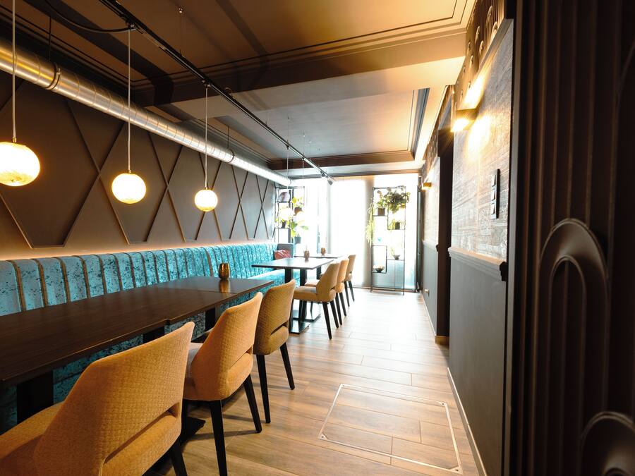 A modern restaurant with a long wooden table, cushioned seating, pendant lighting, and a geometric wall design.