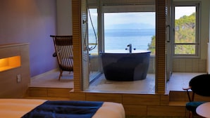 A hotel room with a bed, a chair, a bathtub, and a view of the sea.