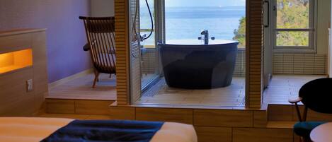 A hotel room with a bed, a chair, a bathtub, and a view of the sea.