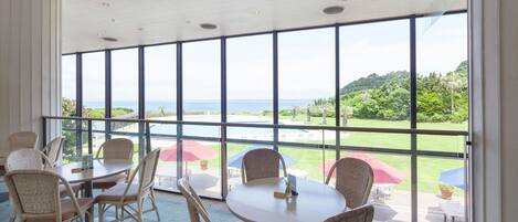 A dining area with wicker chairs and a round table, large windows with a view of a green landscape and the sea.