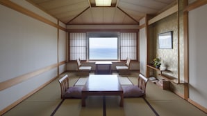 A traditional Japanese room with tatami flooring, a low table, cushions, and a view of the sea through large windows.