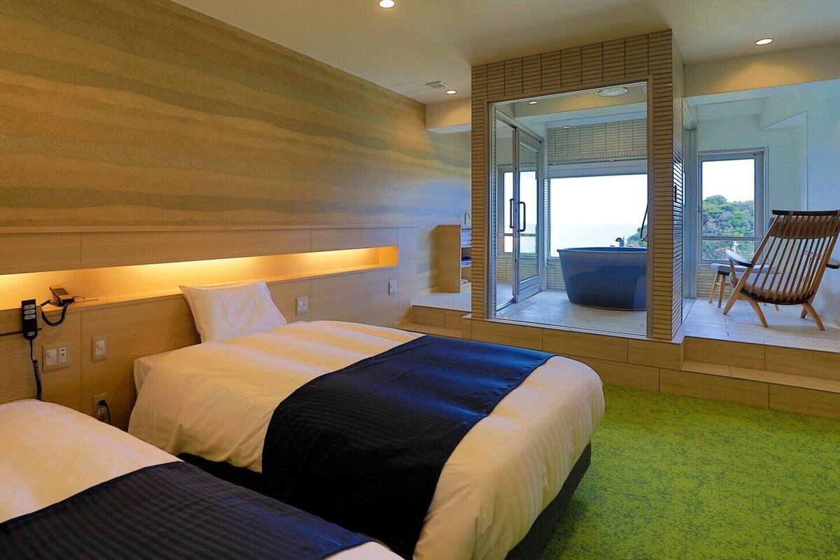 Habitación de hotel con dos camas, un baño con bañera y un balcón con una silla.