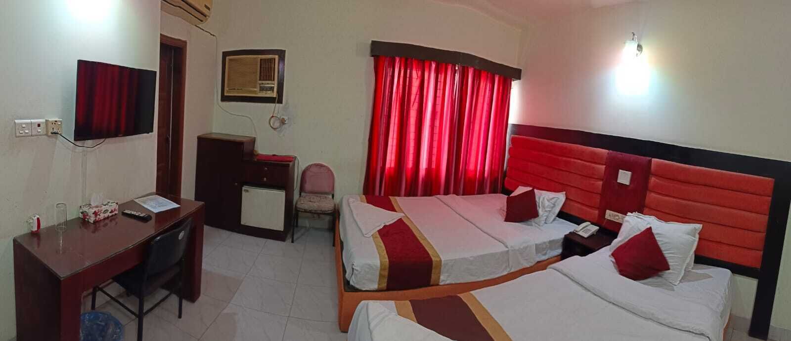 Ein Hotelzimmer mit zwei Betten, einem Schreibtisch, einem Stuhl, einer Schreibtischlampe, einem Radio und einem Fenster mit roten Vorhängen.